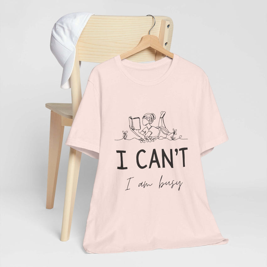 I Can’t, I’m Busy Unisex Jersey Short Sleeve TeeShow off your love for quiet moments with our I Can’t, I’m Busy Unisex Jersey Short Sleeve Tee. Perfect for book lovers and those who cherish their downtime, this tee features a playful illustration of a per